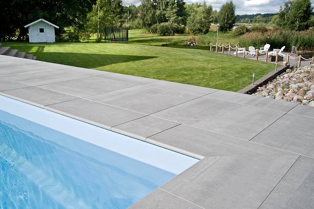 Szare płytki ceramiczne wokół basenu zewnętrznego, z widokiem na trawnik, domek narzędziowy i trampolinę w tle.