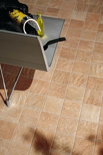 Beżowe płytki podłogowe o nieregularnej fakturze, ułożone w kwadratową siatkę, z metalowym stojakiem na gazety, butelką i czarnym materiałem w rogu.