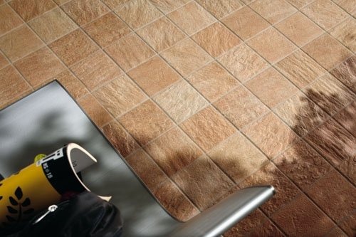 Ułożona pod kątem beżowo-brązowa podłoga z płytek ceramicznych w trakcie prac wykończeniowych, widoczne narzędzia, mata ochronna i częściowo przykryty fragment podłogi z różnicą w odcieniu płytek.