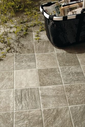 Szare, kwadratowe płytki podłogowe z teksturą kamienia, koszyk z gazetami i zielone pnącze w rogu kadru.