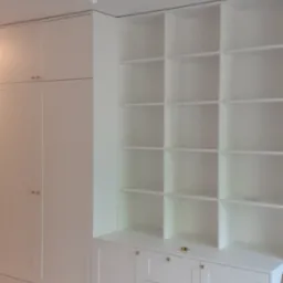 Biała zabudowa meblowa z szafami i regałem z półkami, widoczne złote uchwyty i narzędzie na blacie, minimalistyczny design, jasne oświetlenie.