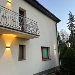 Sebastiansak - Elewacja domu z zamontowanym oświetleniem zewnętrznym: dwa kinkiety na parterze i jeden pod balkonem, ozdobna balustrada balkonowa, okna z brązowymi ramami, iglaki w ogrodzie.