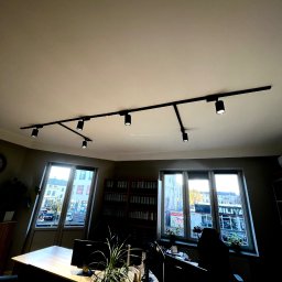 Sebastiansak - Biuro z nowoczesnym oświetleniem szynowym, widok na ulicę przez okna, biurko z komputerami i rośliną doniczkową.