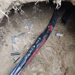 Sebastiansak - Połączenie kabli elektrycznych w wykopie, widoczna izolacja z taśmą w kolorze czerwonym i niebieskim, obok leżą fragmenty izolacji, stopa w gumowym bucie w błocie.