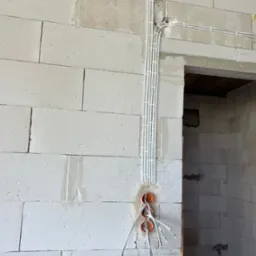 Surowe wnętrze w trakcie prac: widoczne przewody elektryczne w białych rurkach przymocowanych do ściany z betonu komórkowego, wystające z pomarańczowych puszek instalacyjnych.