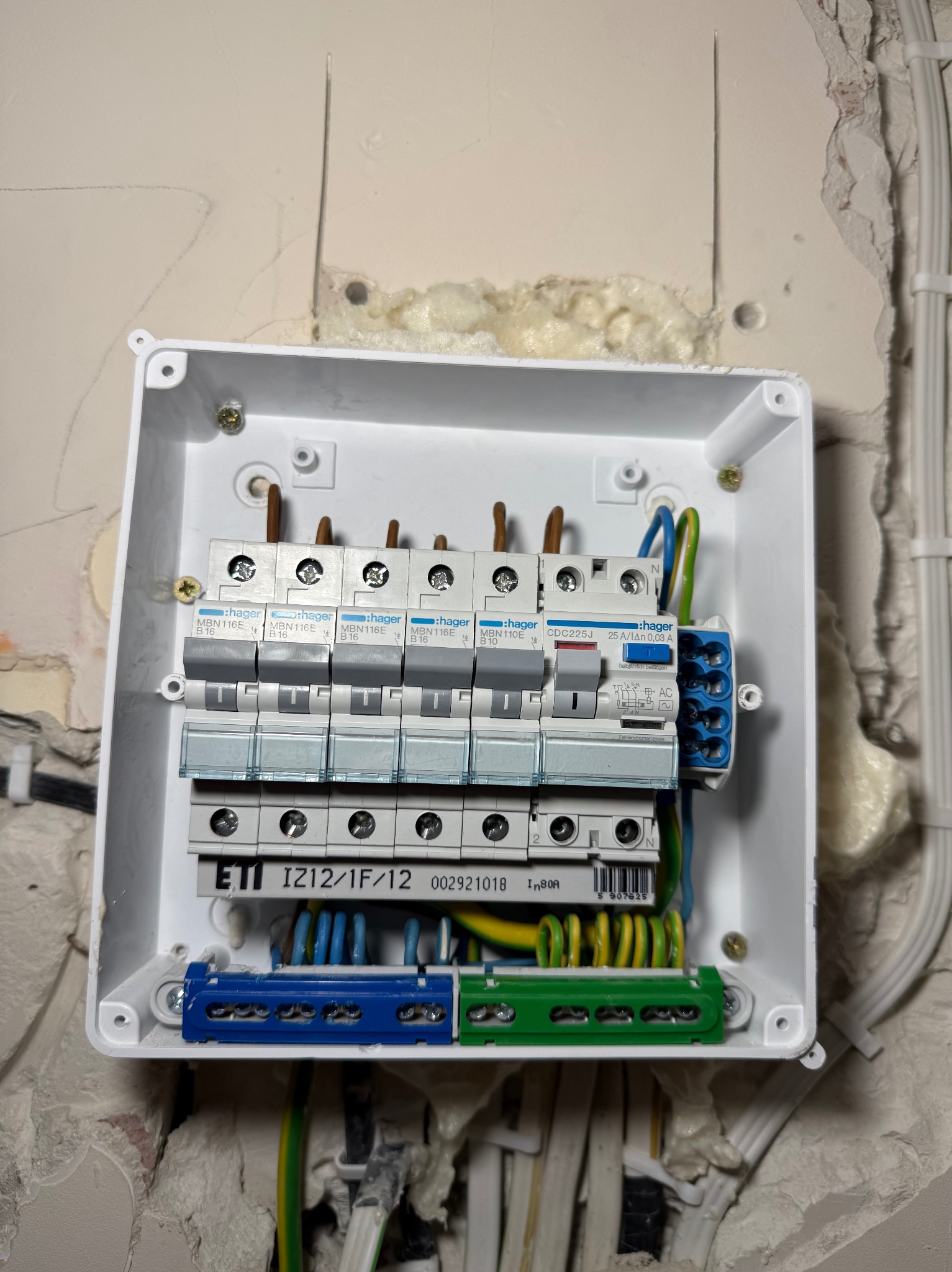 Otwarta skrzynka elektryczna z zamontowanymi bezpiecznikami Hager MBN116E, zabezpieczeniem różnicowoprądowym Hager CDC225J i listwami zaciskowymi ETI IZ12/1F/12, widoczne kable w kolorach...