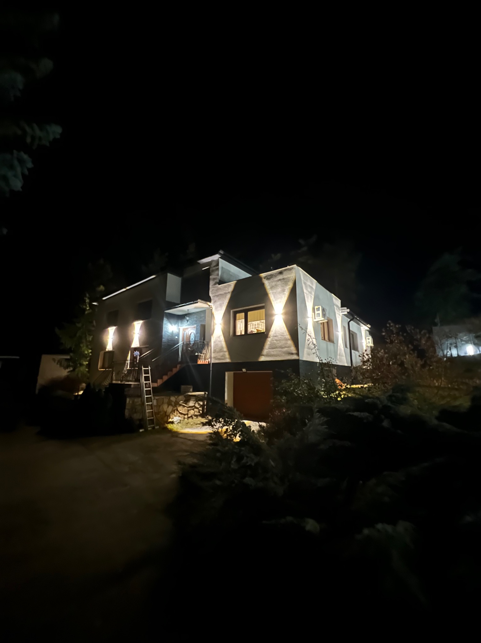 Nowoczesny dom jednorodzinny oświetlony zewnętrznymi lampami ściennymi w kształcie diamentów, widoczna drabina oparta o schody wejściowe, nocne ujęcie.