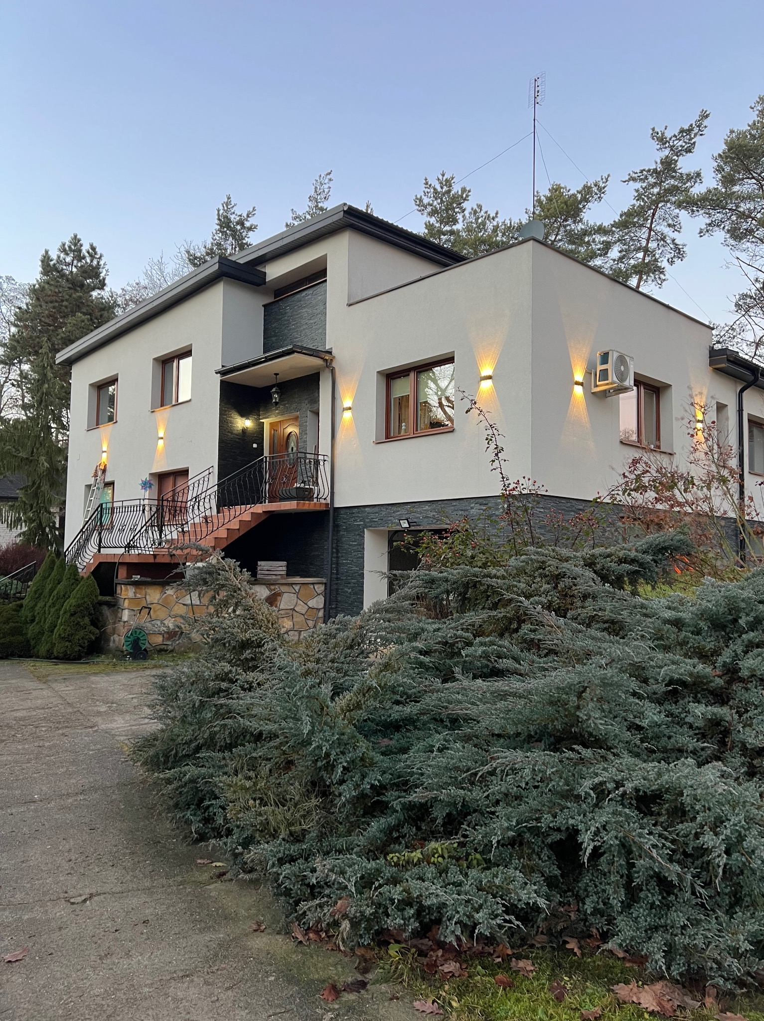 Elewacja domu z oświetleniem zewnętrznym, schodami z kutą balustradą i klimatyzatorem na ścianie.