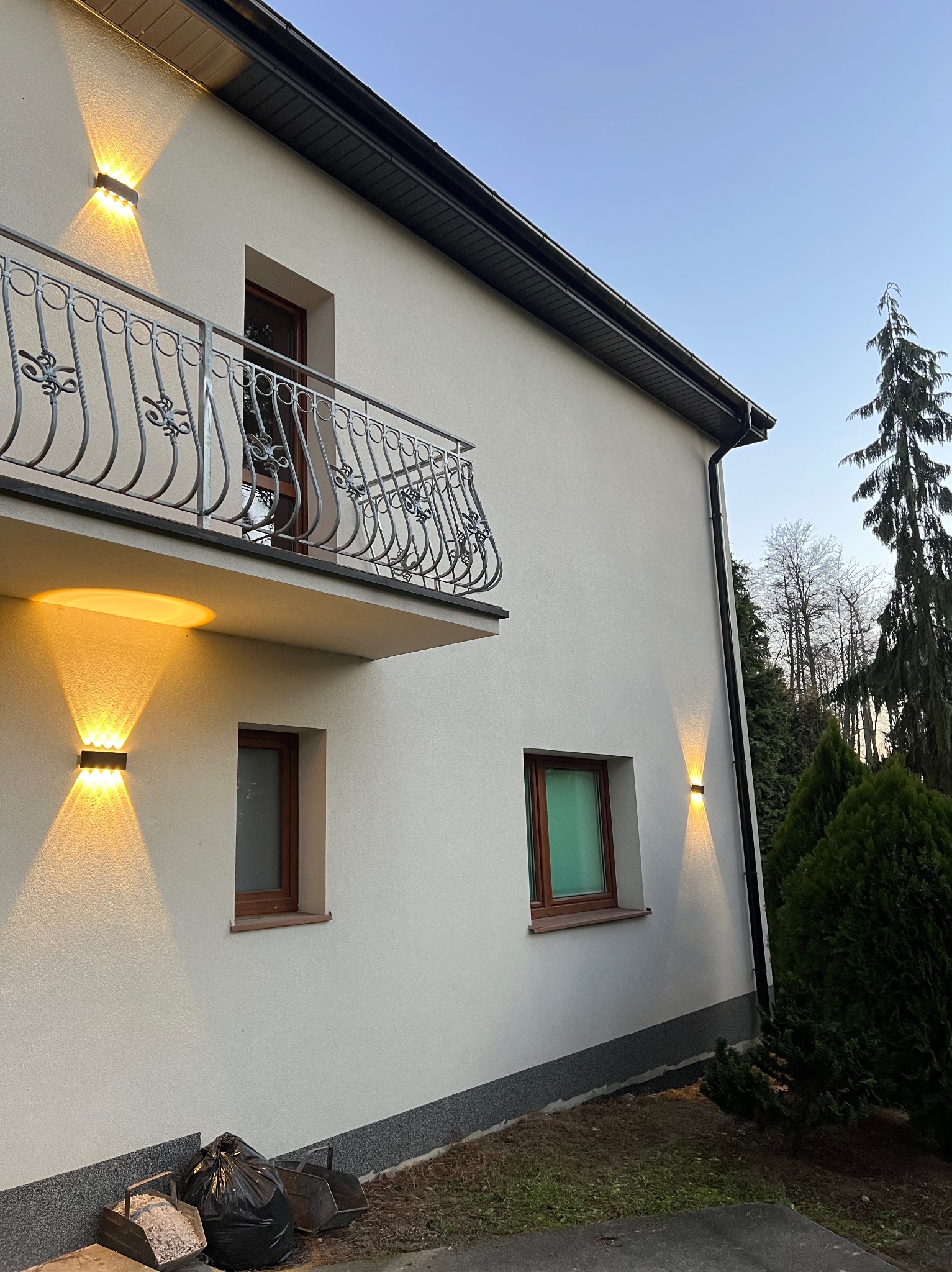 Elewacja domu z zamontowanym oświetleniem zewnętrznym: dwa kinkiety na parterze i jeden pod balkonem, ozdobna balustrada balkonowa, okna z brązowymi ramami, iglaki w ogrodzie.