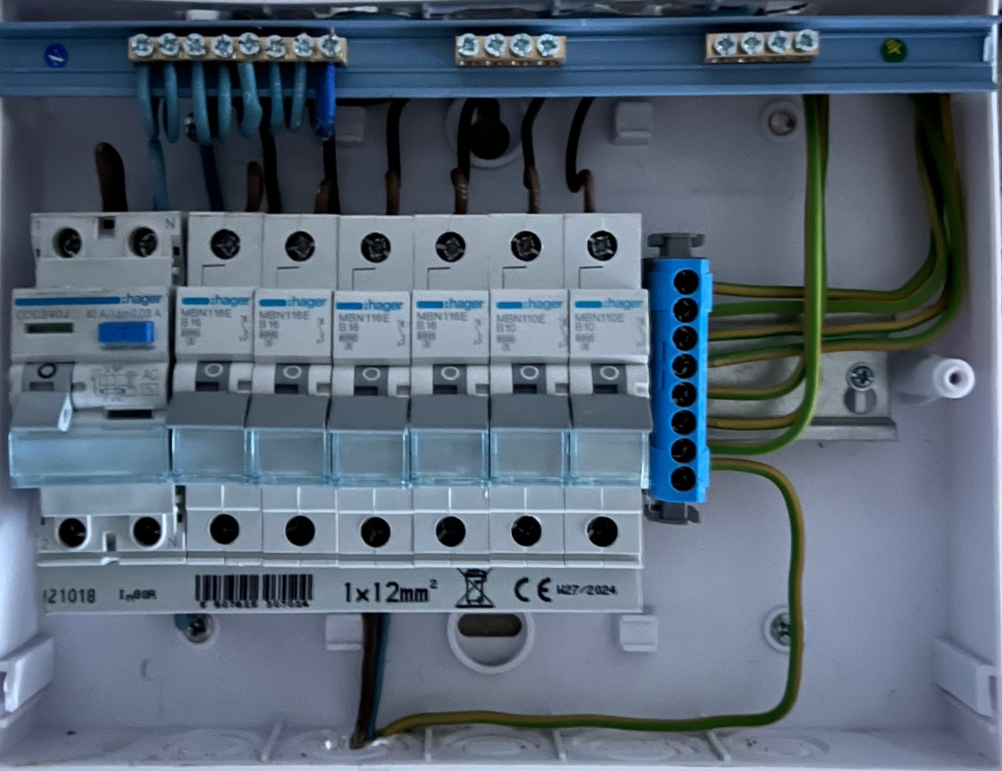 Wewnętrzny widok skrzynki rozdzielczej z zainstalowanymi bezpiecznikami Hager, niebieską listwą zaciskową i przewodami w kolorach brązowym, niebieskim i zielono-żółtym.