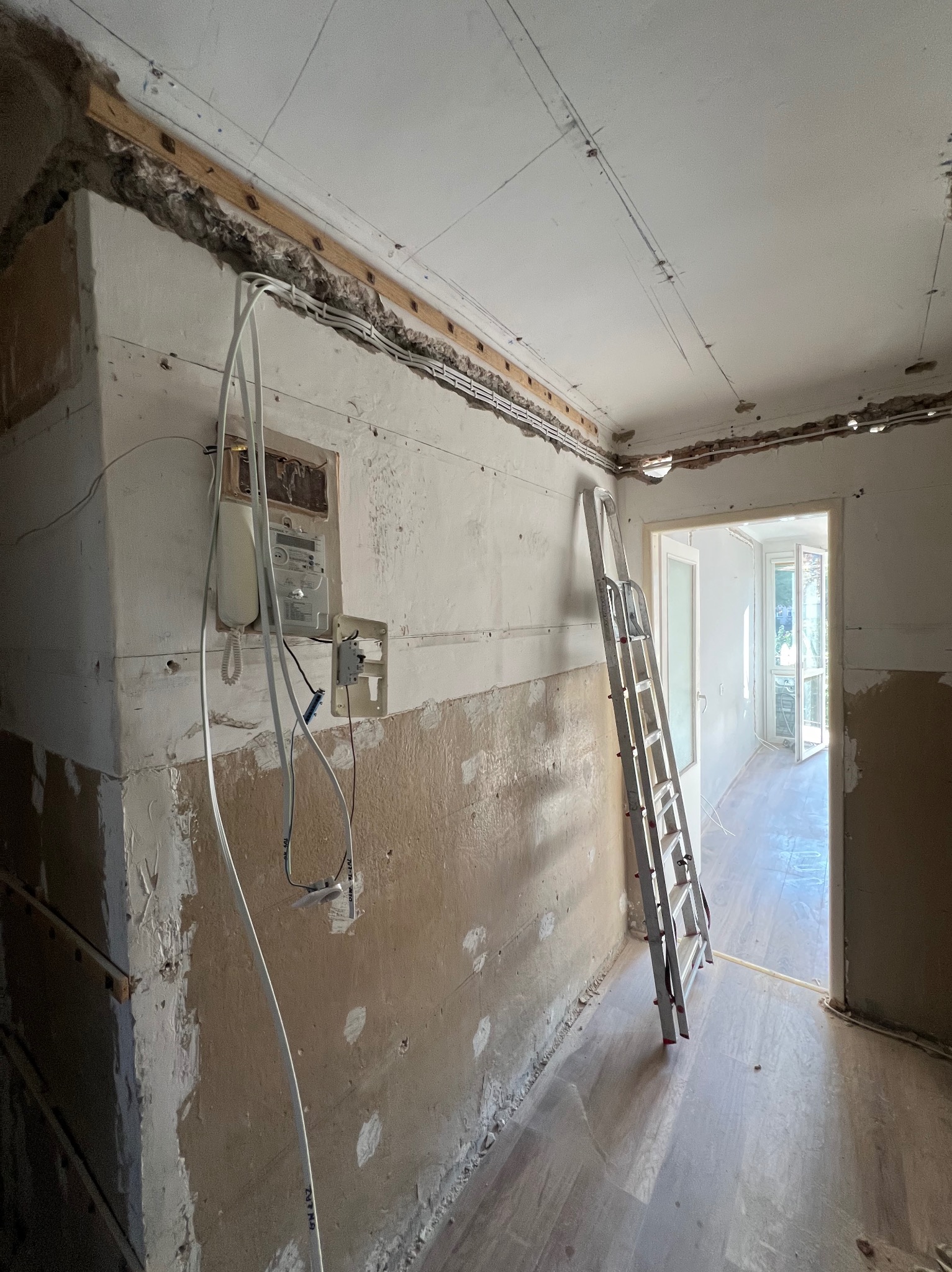 Wnętrze w trakcie remontu: odsłonięte przewody elektryczne, licznik, fragmenty starej farby na ścianach, aluminiowa drabina oparta o ścianę, widok na sąsiednie pomieszczenie z jasną podłogą.