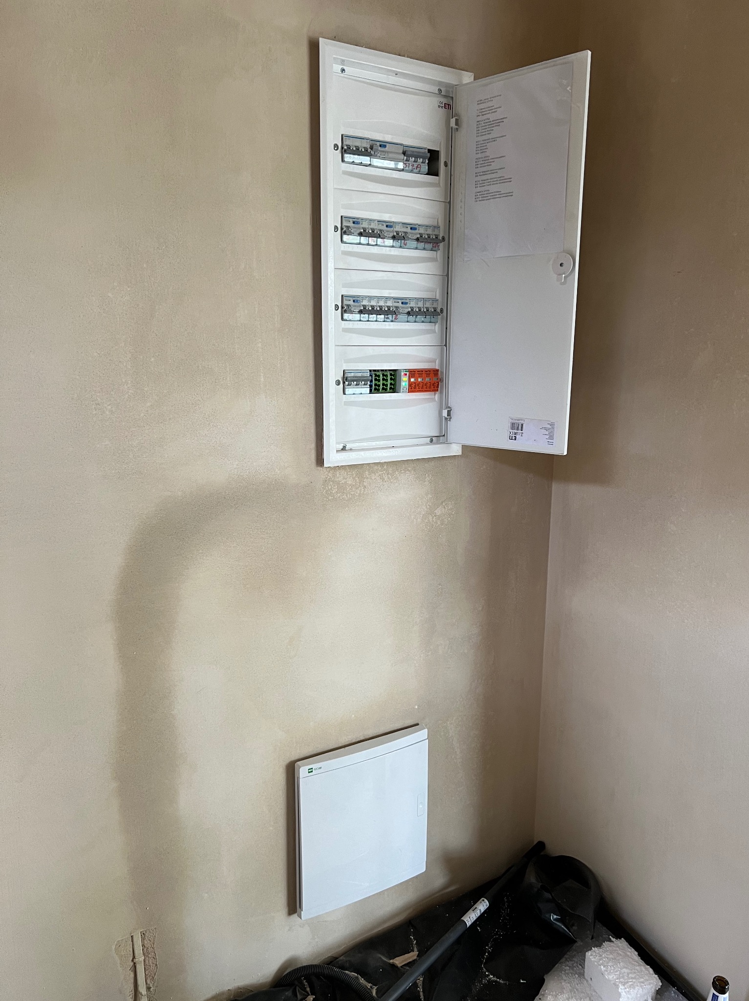 Rozdzielnica elektryczna z otwartymi drzwiczkami na ścianie w surowym stanie, poniżej zamontowany grzejnik elektryczny.
