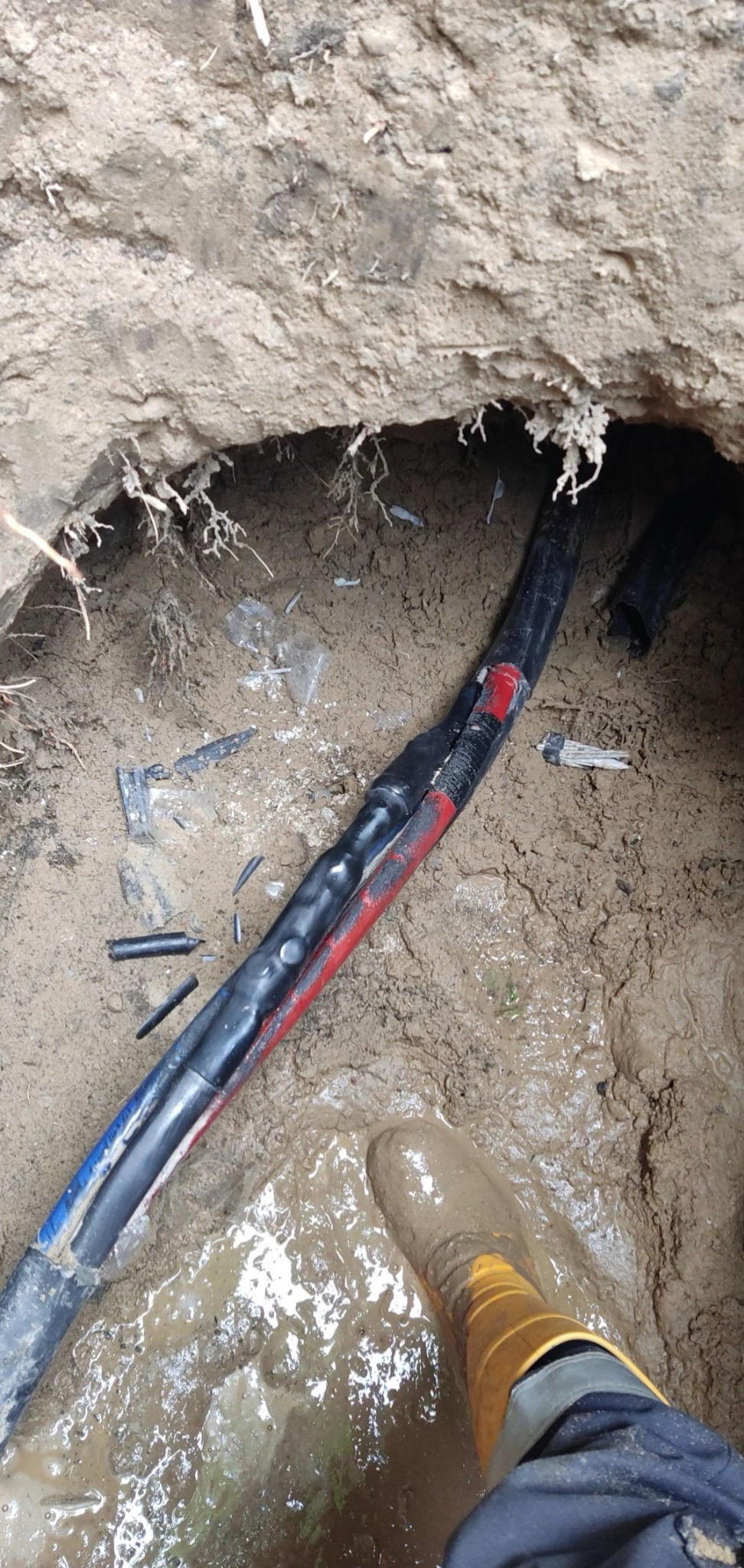 Połączenie kabli elektrycznych w wykopie, widoczna izolacja z taśmą w kolorze czerwonym i niebieskim, obok leżą fragmenty izolacji, stopa w gumowym bucie w błocie.