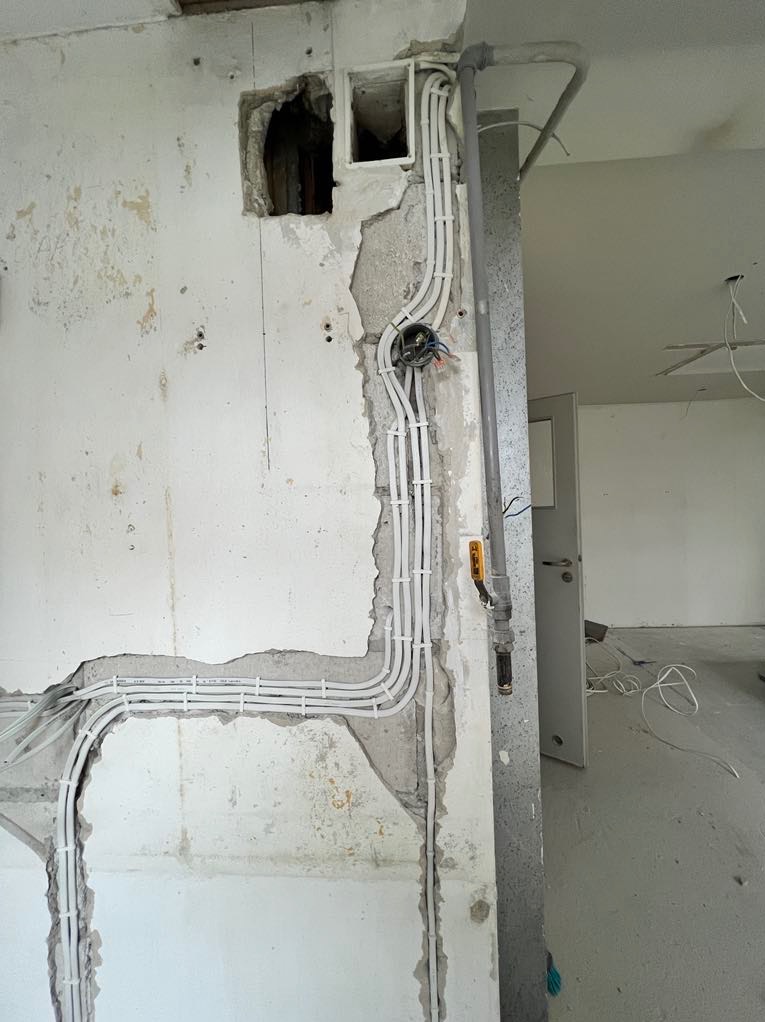 Ściana w trakcie remontu z nową instalacją elektryczną: białe kable prowadzone wzdłuż ściany, puszka elektryczna z widocznymi przewodami, fragment otworu w ścianie, szare rury pionowe.