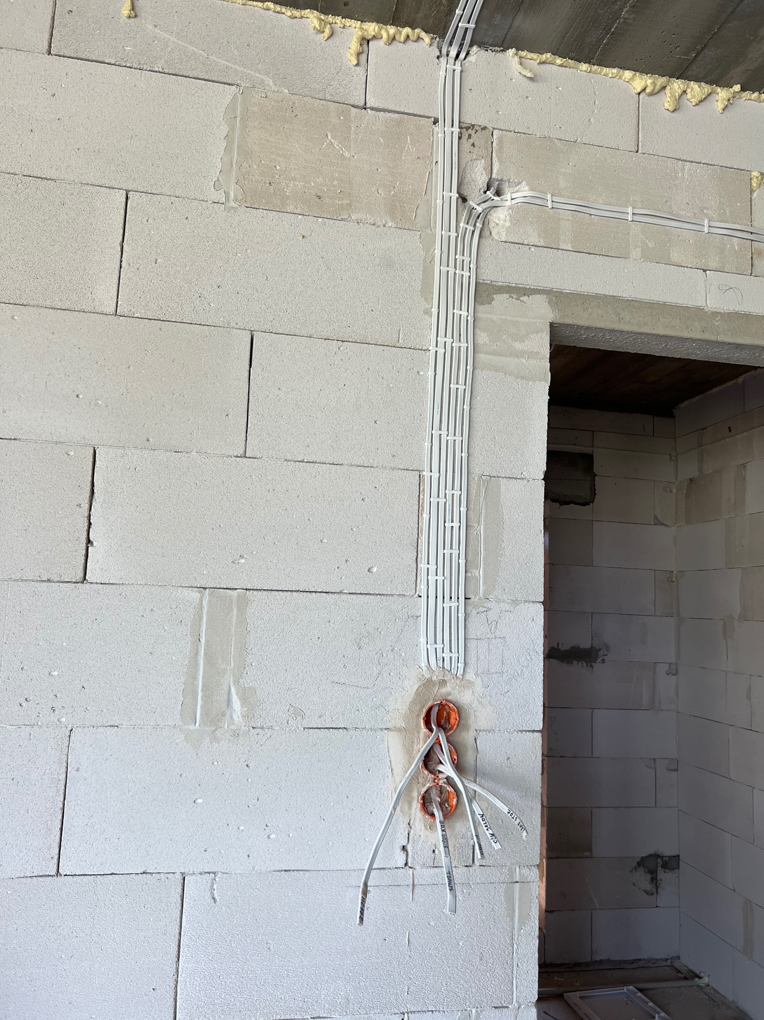 Surowe wnętrze w trakcie prac: widoczne przewody elektryczne w białych rurkach przymocowanych do ściany z betonu komórkowego, wystające z pomarańczowych puszek instalacyjnych.