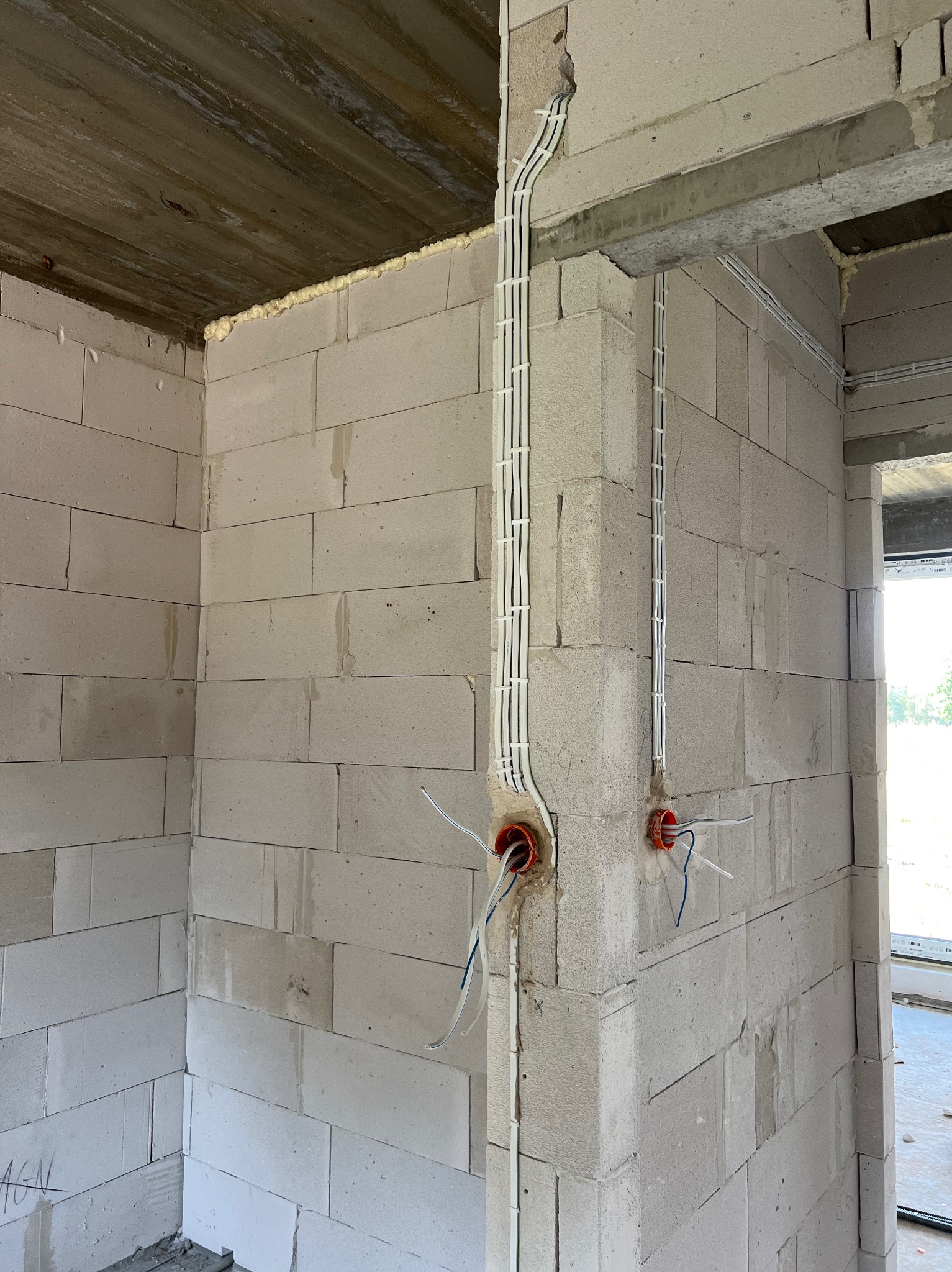 Wewnętrzne ściany z betonu komórkowego w surowym stanie z widocznymi, pionowo poprowadzonymi przewodami elektrycznymi w białych korytkach; widoczne puszki instalacyjne z wystającymi przewodami.