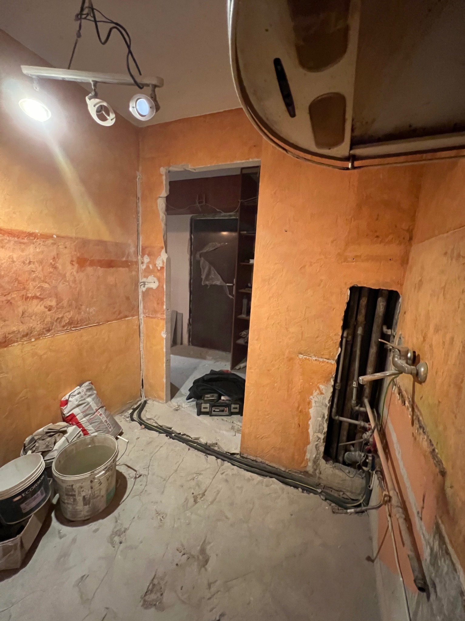 Pomarańczowe ściany w trakcie remontu, widoczne odsłonięte rury, przewody elektryczne, wiadro z wodą i worki z materiałami budowlanymi, oświetlenie sufitowe.