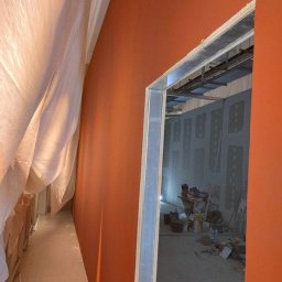 Firma budowlana WBR - Pomarańczowa ściana w trakcie malowania, widok przez metalową ramę drzwi do pomieszczenia z płytami kartonowo-gipsowymi i sprzętem budowlanym, zabezpieczona folią podłoga.