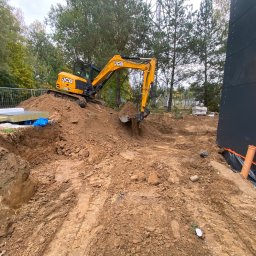 Firma budowlana WBR - Żółta koparka JCB wykopuje ziemię pod budowę, widoczne materiały izolacyjne i fragmenty rur kanalizacyjnych, w tle drzewa i element konstrukcji stalowej