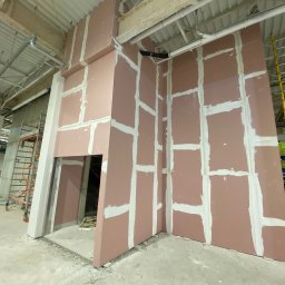 Firma budowlana WBR - Montaż różowych płyt gipsowo-kartonowych na metalowym stelażu wewnątrz budynku, widoczne białe spoiny, surowy beton na podłodze i drabiny w tle.