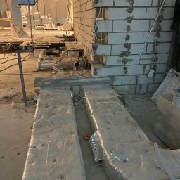 Firma budowlana WBR - Surowy stan wnętrza w trakcie budowy: widoczne przewody wentylacyjne owinięte folią aluminiową, ściana z pustaków oraz liczne kable i rury pod sufitem.