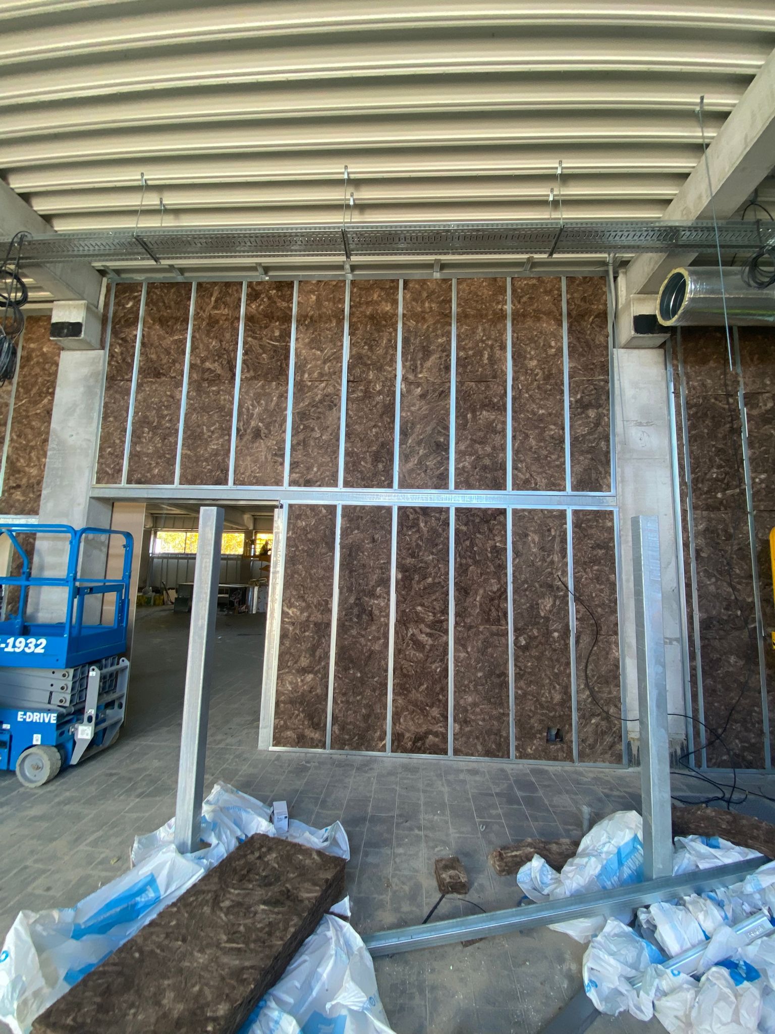 Montaż izolacji z wełny mineralnej w stalowym stelażu w przestrzeni biurowej, widoczny podnośnik koszowy i materiały budowlane na podłodze.