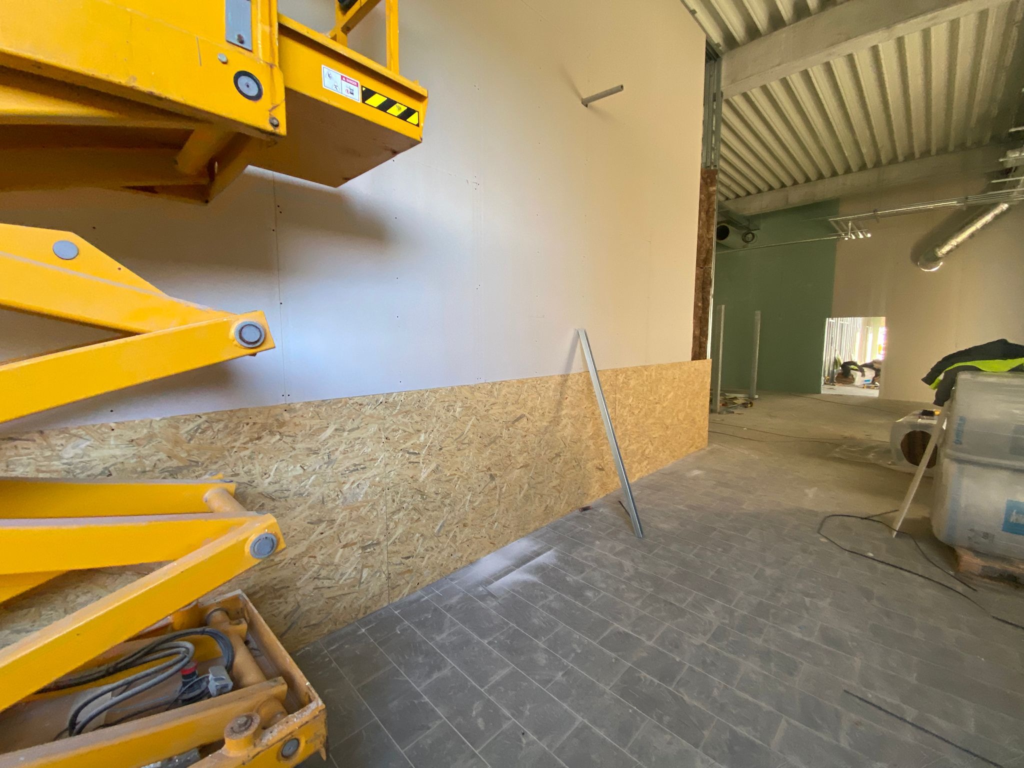 Wnętrze biura w trakcie remontu: częściowo wykończona ściana z płyt OSB i gipsowo-kartonowych, widoczny podnośnik nożycowy i elementy instalacji pod sufitem.