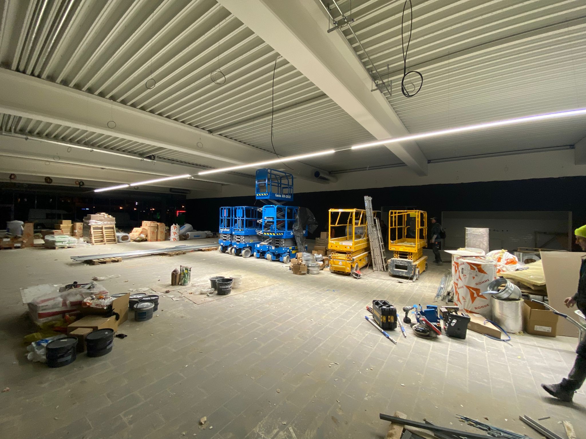 Wnętrze remontowanego biura z widocznym surowym sufitem, podnośnikami koszowymi Genie GS-1932 w kolorach niebieskim i żółtym, materiałami budowlanymi oraz narzędziami, przygotowane do montażu płyty...