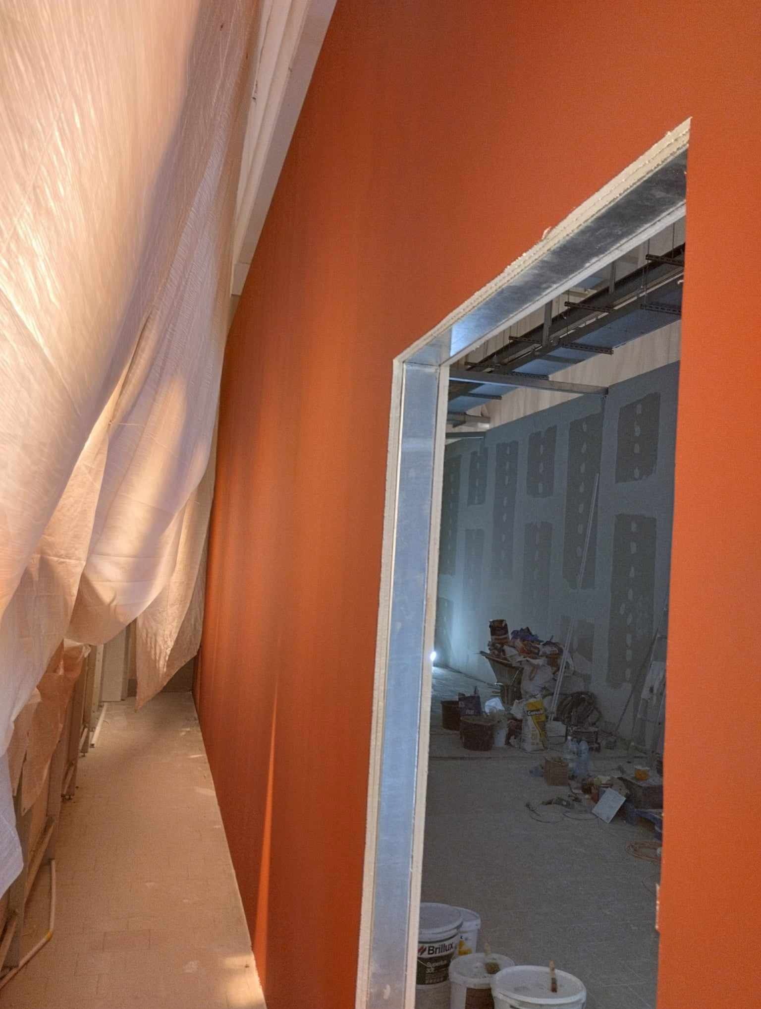 Pomarańczowa ściana w trakcie malowania, widok przez metalową ramę drzwi do pomieszczenia z płytami kartonowo-gipsowymi i sprzętem budowlanym, zabezpieczona folią podłoga.
