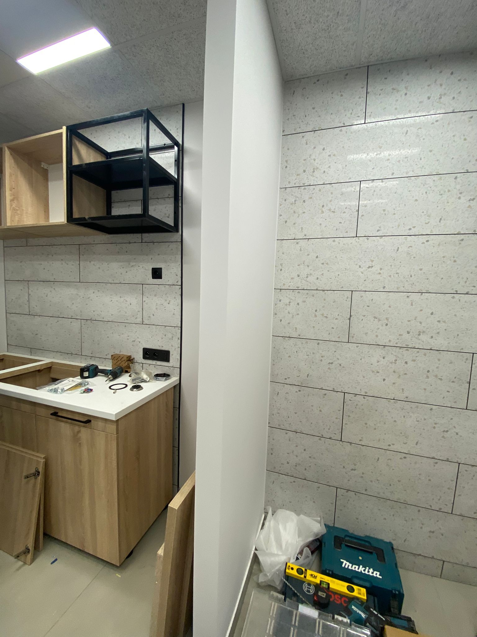 Wnętrze w trakcie montażu: fragment szafki kuchennej z białym blatem, czarna metalowa półka wisząca, płytki imitujące kamień na ścianie, narzędzia na blacie, skrzynka narzędziowa Makita na podłodze.