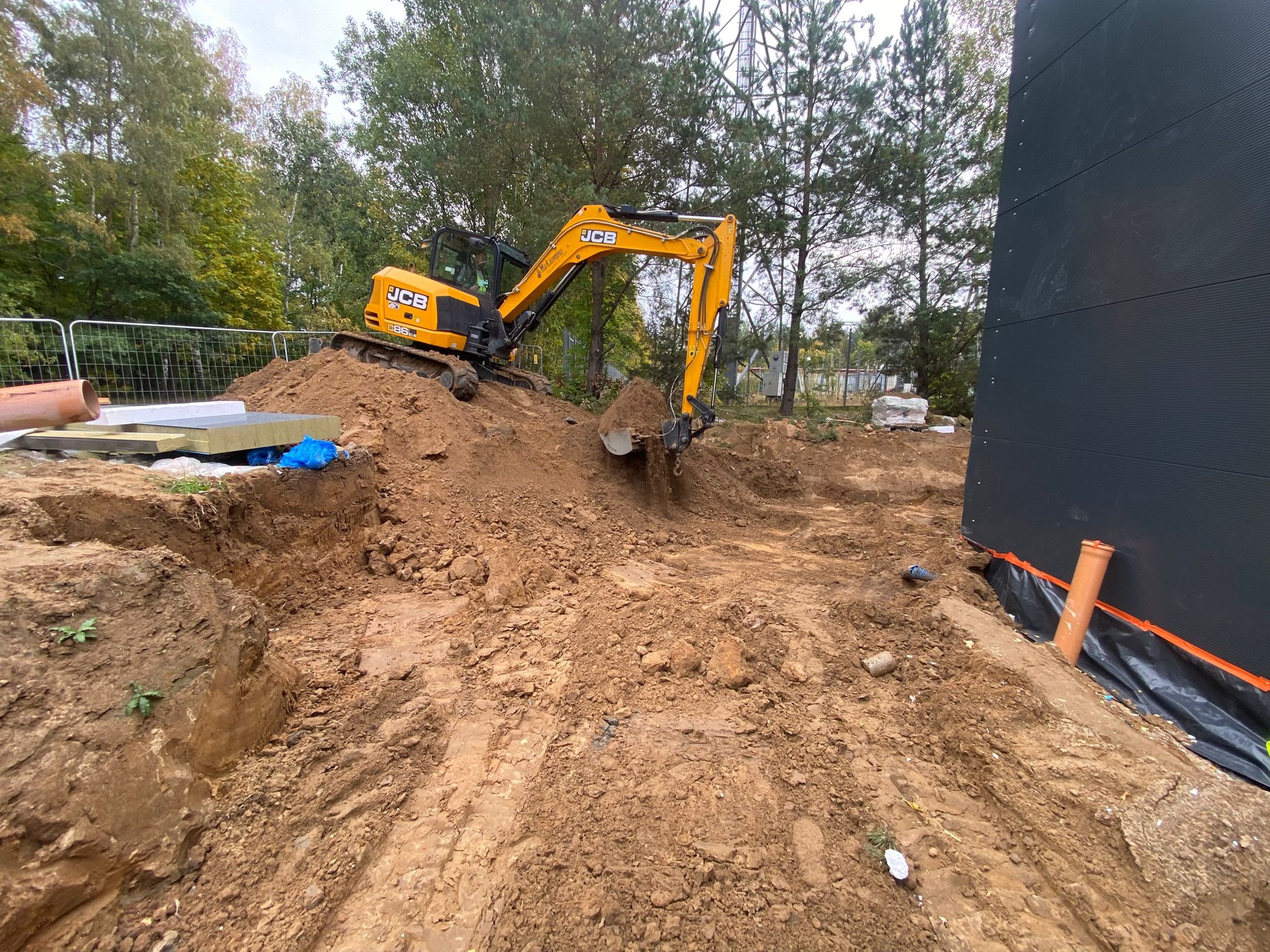 Żółta koparka JCB wykopuje ziemię pod budowę, widoczne materiały izolacyjne i fragmenty rur kanalizacyjnych, w tle drzewa i element konstrukcji stalowej