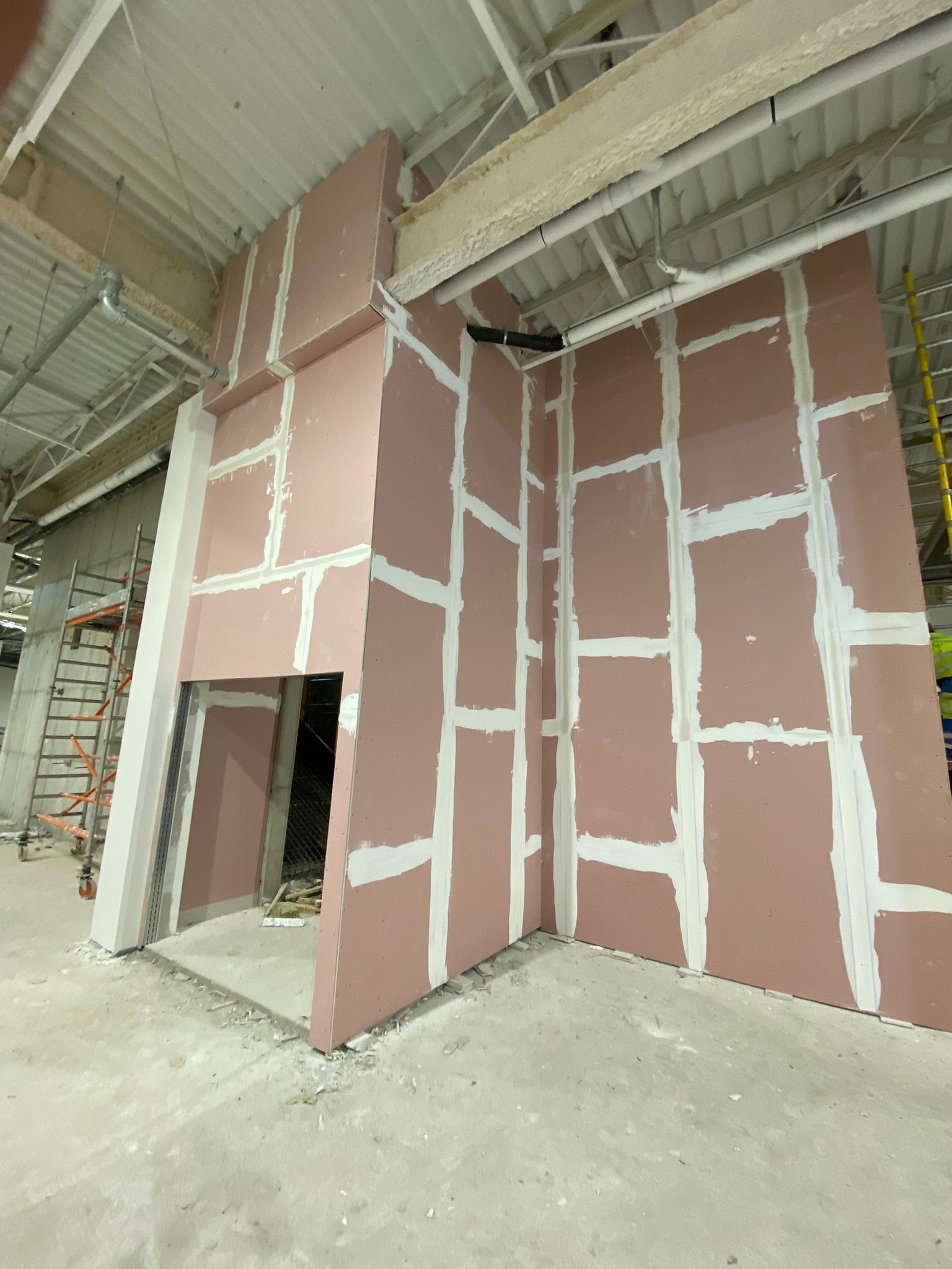 Montaż różowych płyt gipsowo-kartonowych na metalowym stelażu wewnątrz budynku, widoczne białe spoiny, surowy beton na podłodze i drabiny w tle.