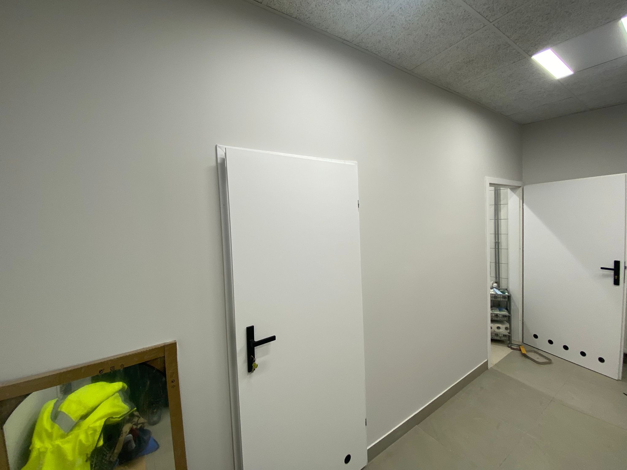 Nowo pomalowane, jasnoszare ściany biura z zamontowanymi, białymi drzwiami z czarnymi klamkami, widok na korytarz i pomieszczenie gospodarcze z regałem.