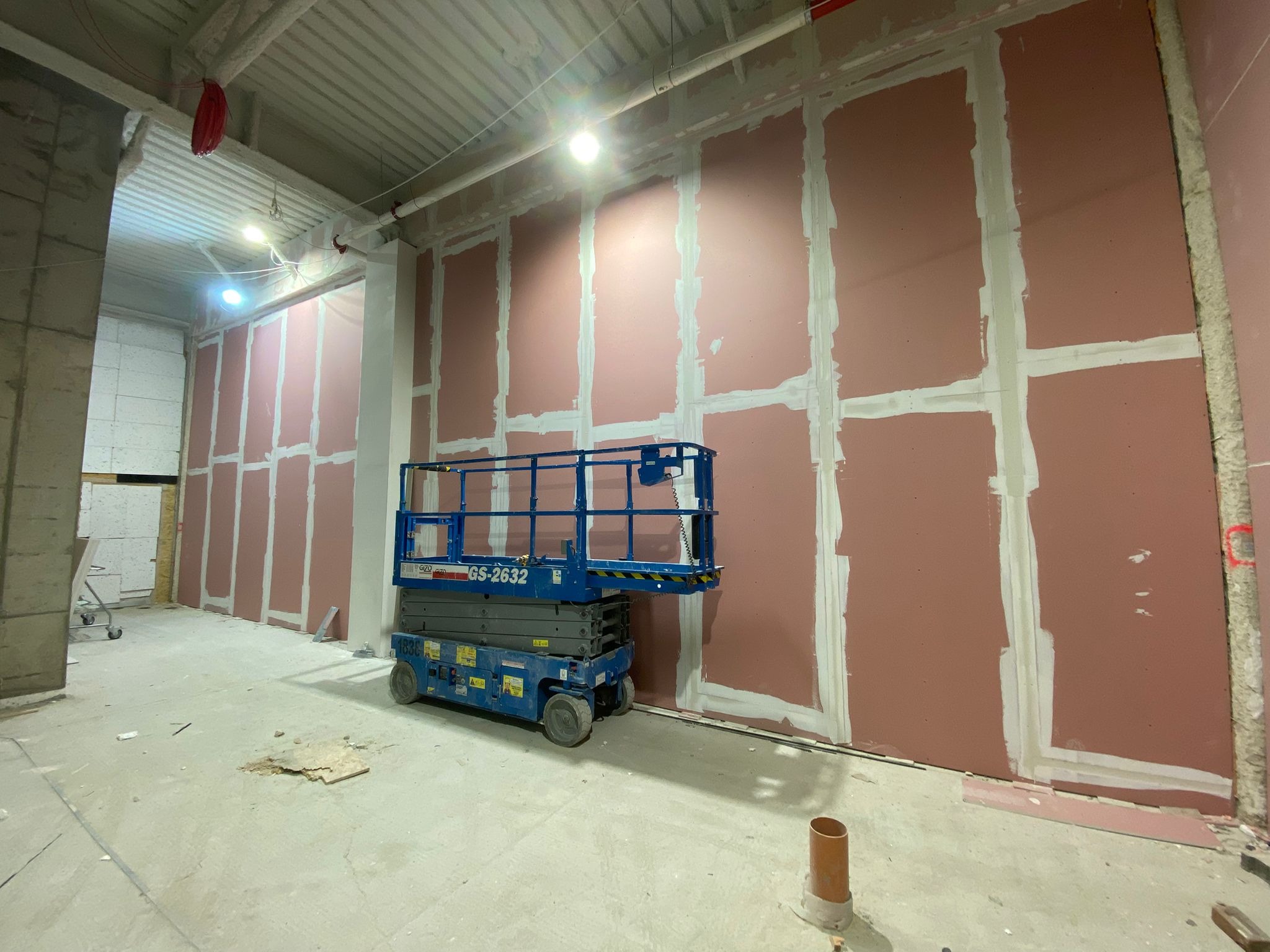 Montaż płyt karton gips w biurze, widoczna ściana z płytami w kolorze różowym, szpachlowane łączenia, podnośnik nożycowy niebieski, surowy stan wykończenia