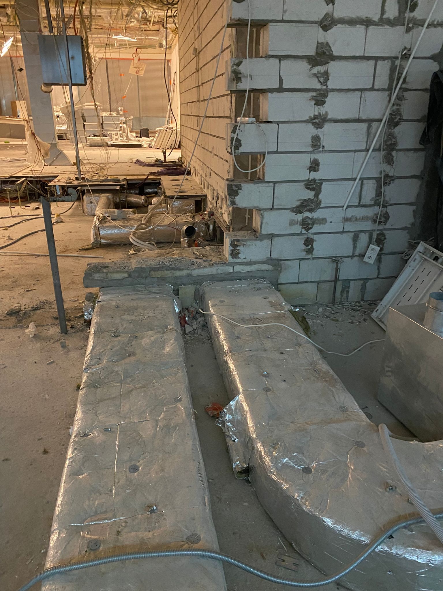 Surowy stan wnętrza w trakcie budowy: widoczne przewody wentylacyjne owinięte folią aluminiową, ściana z pustaków oraz liczne kable i rury pod sufitem.