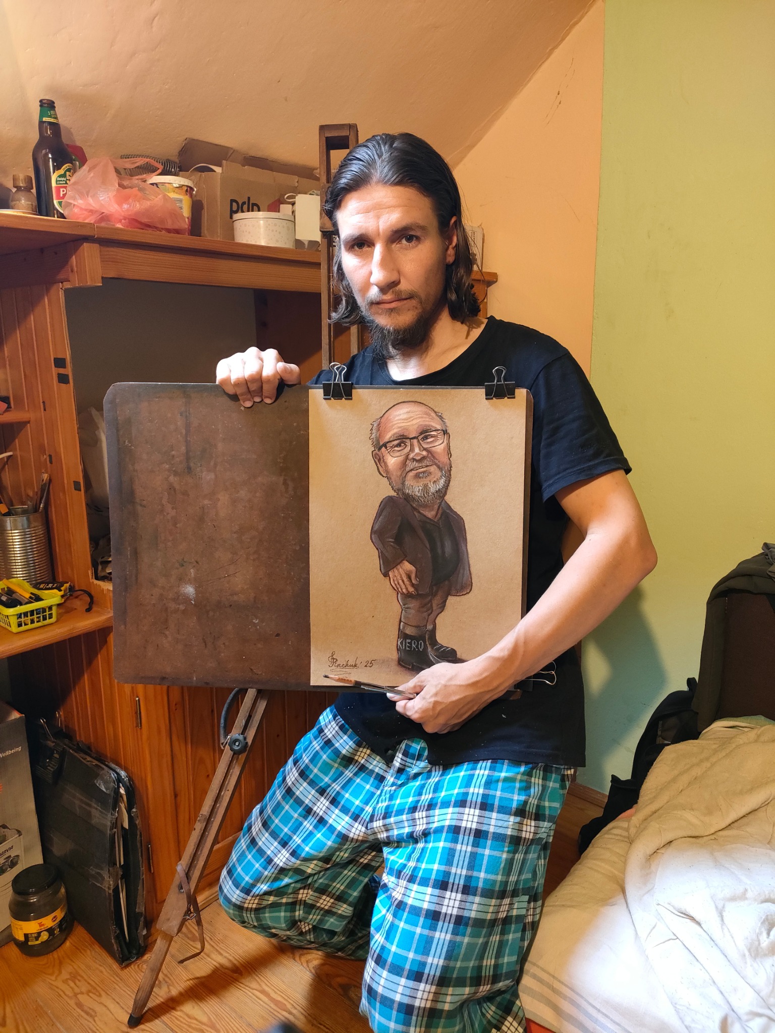 Artysta z Kazimierza Dolnego prezentuje karykaturę na brązowym papierze, przypiętą do deski klipsami. Widoczny podpis autora i data. Ujęcie z bliska, w domowym otoczeniu.