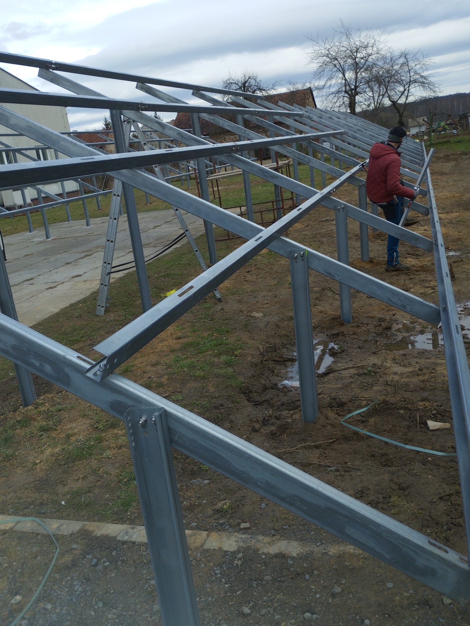 Konstrukcja szkieletowa z metalowych profili, prawdopodobnie pod budynek gospodarczy lub szklarnię, z pracującym mężczyzną w czerwonej kurtce.