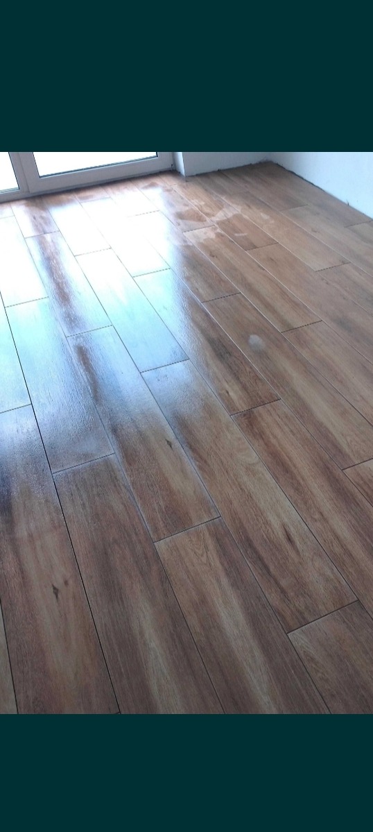 Ułożona pod kątem, brązowa podłoga z płytek imitujących drewno, z widocznym oknem w tle.