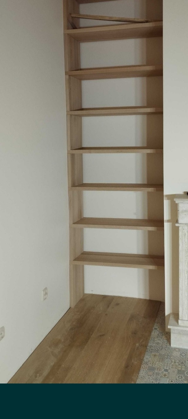 Wnęka z drewnianymi półkami w kolorze jasnego dębu, zamontowana między ścianą a elementem dekoracyjnym przypominającym kominek, z widoczną podłogą z paneli i fragmentem wzorzystej posadzki.