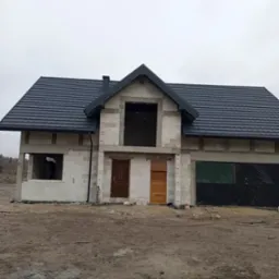 Dom w stanie surowym zamkniętym z nowym, ciemnym dachem z dachówki ceramicznej, widoczne obróbki blacharskie i komin, na zewnątrz teren budowy.