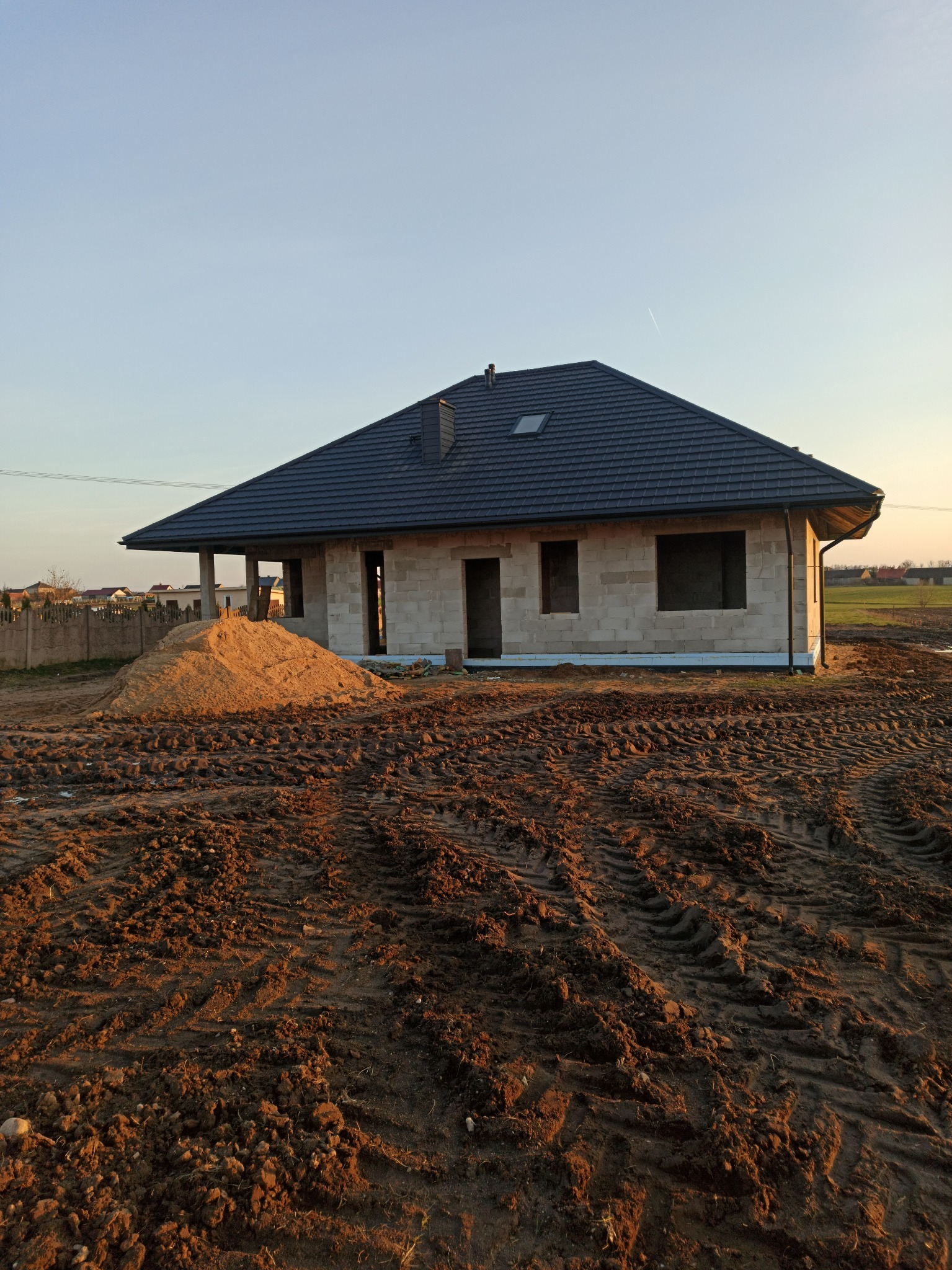 Dom w trakcie budowy z ciemnoszarą dachówką, widoczne ślady opon na ziemi i sterta piasku obok budynku.