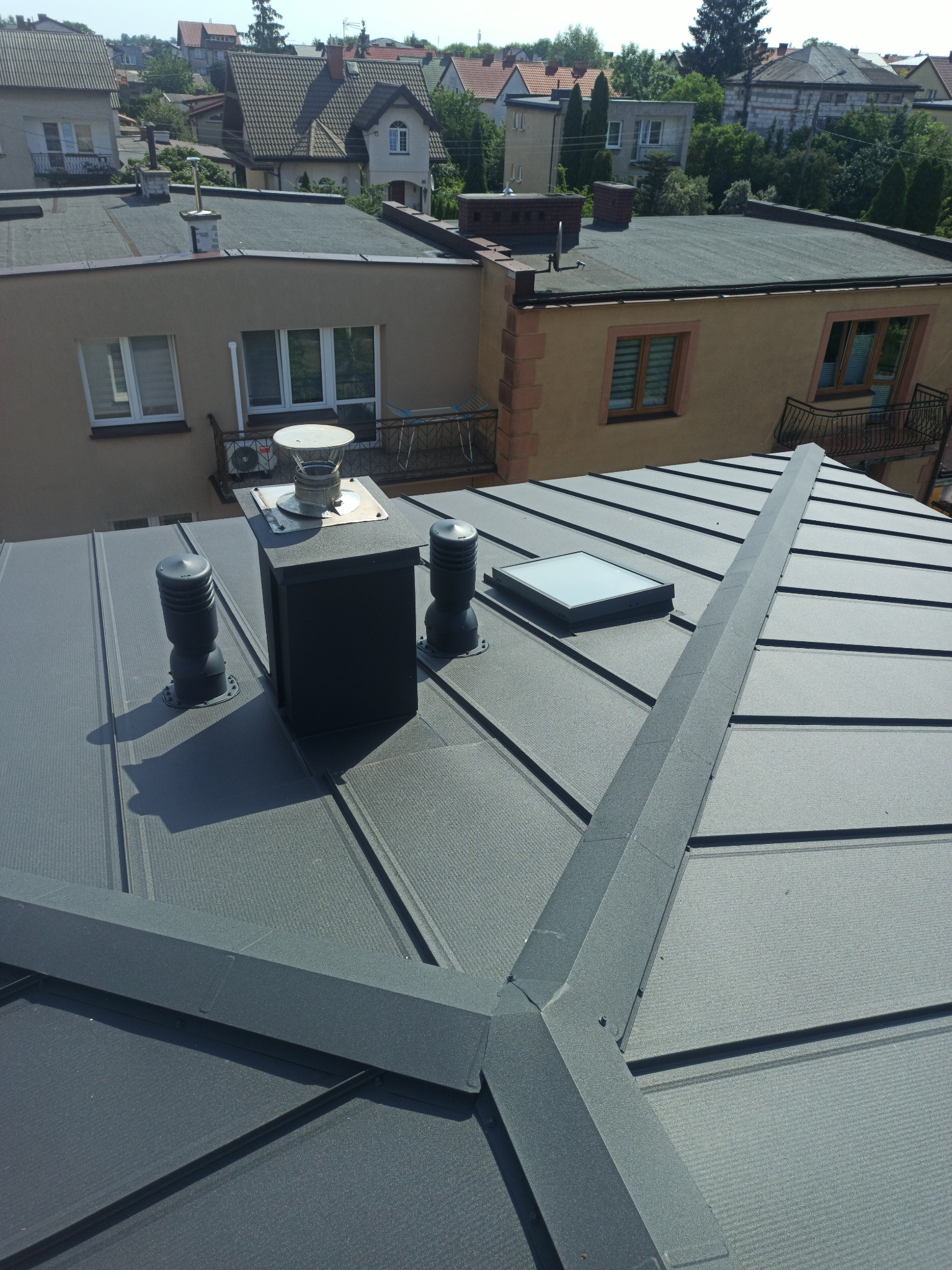 Szary dach z blachy na budynku mieszkalnym, widoczne kominy wentylacyjne, okno dachowe i listwa kalenicowa, w tle zabudowa jednorodzinna.