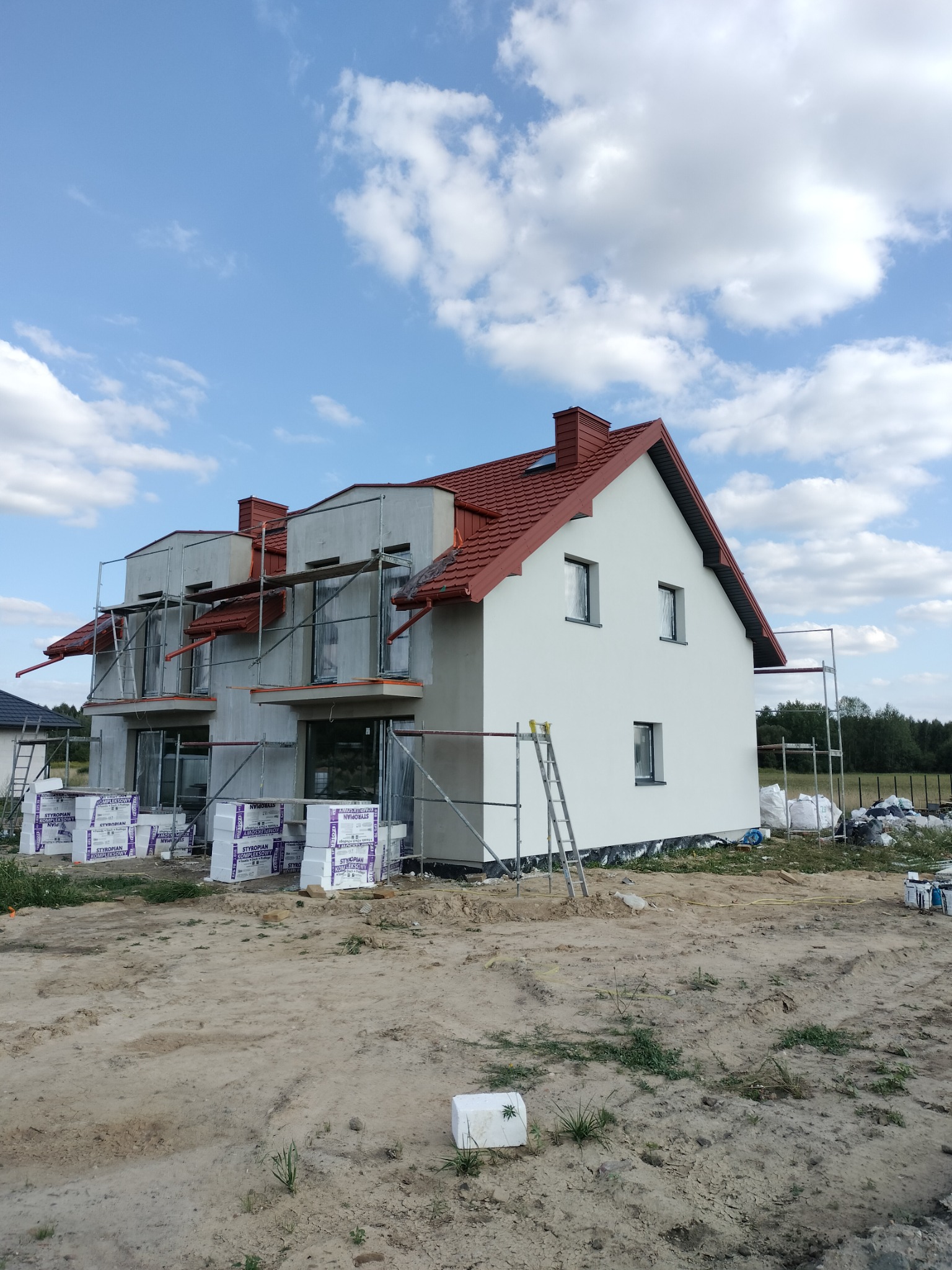 Budynek bliźniaczy w trakcie budowy z częściowo wykonaną elewacją i czerwoną dachówką, otoczony rusztowaniami i materiałami budowlanymi na piaszczystym terenie.