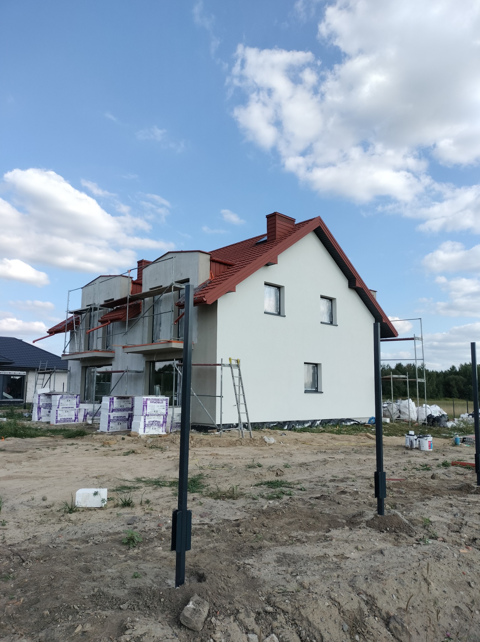 Nowy dom w trakcie budowy z czerwoną dachówką, widoczne rusztowania i materiały izolacyjne na placu budowy w słoneczny dzień.