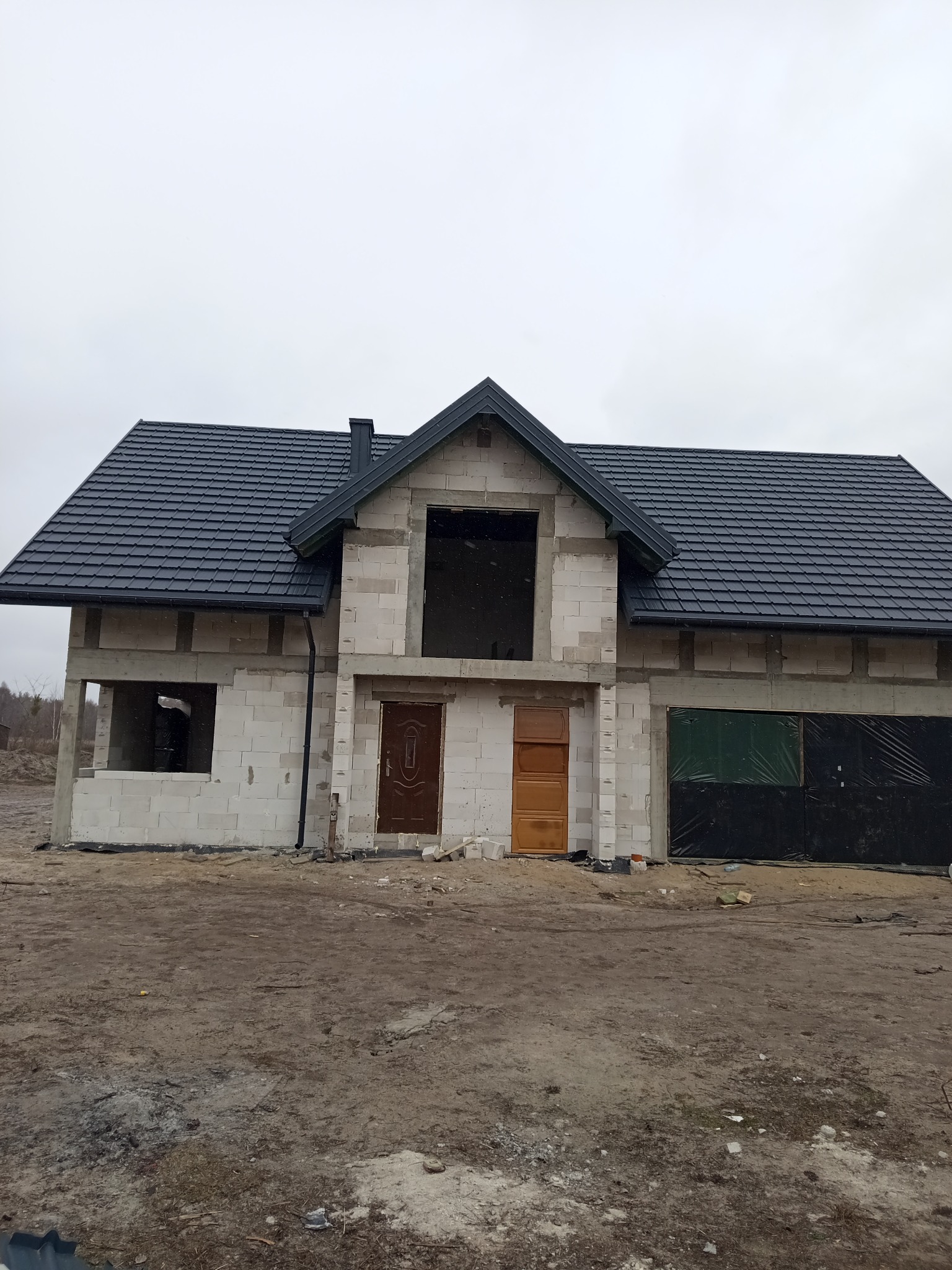 Dom w stanie surowym zamkniętym z nowym, ciemnym dachem z dachówki ceramicznej, widoczne obróbki blacharskie i komin, na zewnątrz teren budowy.