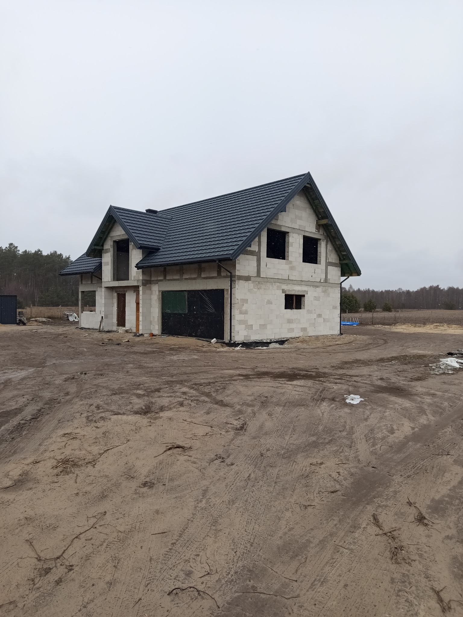 Surowy stan domu jednorodzinnego w trakcie budowy z ciemną dachówką na dachu, widoczne niezabezpieczone otwory okienne i drzwiowe, ziemia wokół rozjeżdżona przez ciężki sprzęt.