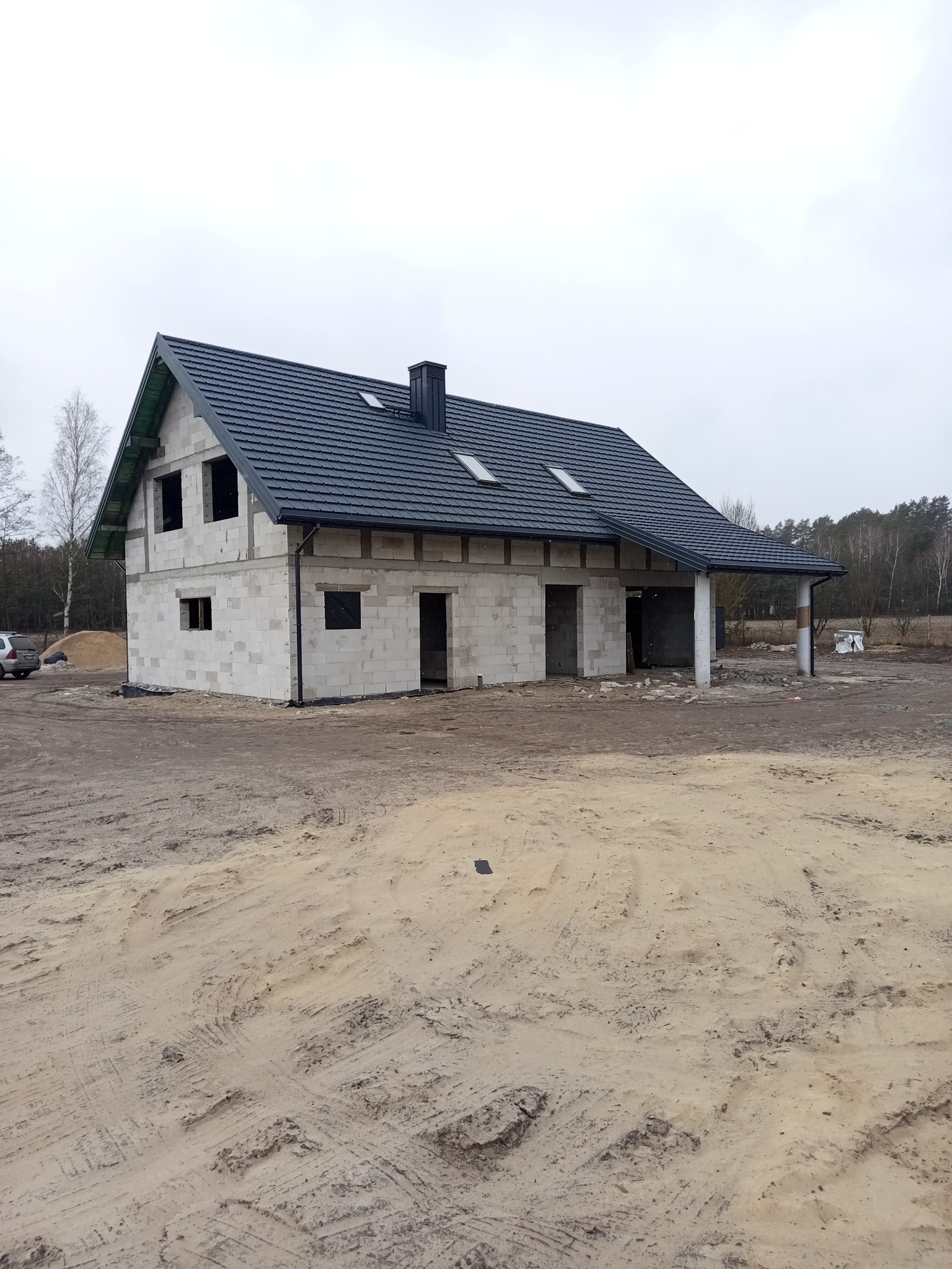 Dom w trakcie budowy z ciemnym dachem, widoczne okna dachowe i komin, surowy teren wokół.