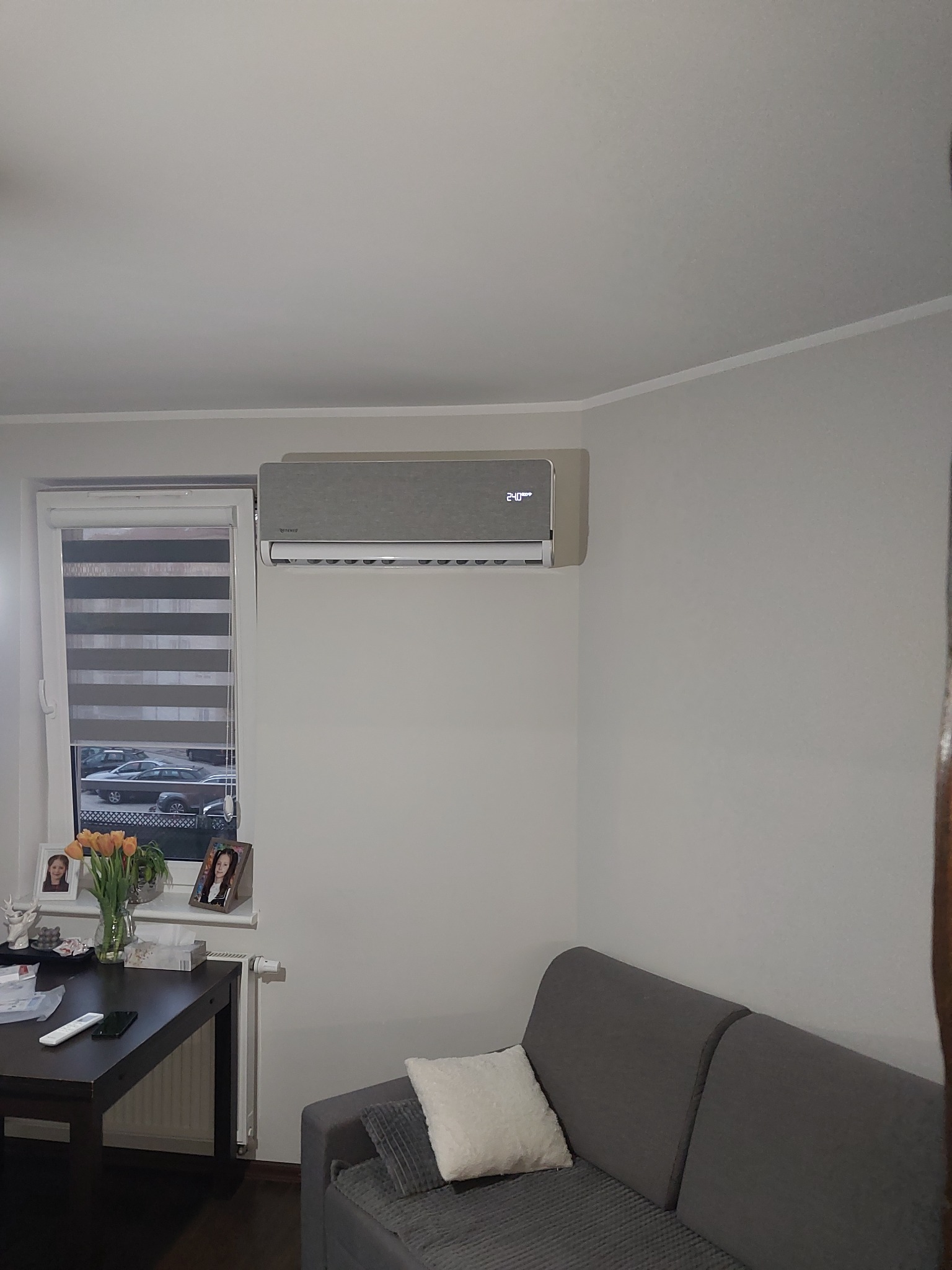 Szary klimatyzator ścienny w salonie, widoczny wyświetlacz temperatury, obok okno z roletami i dekoracjami na parapecie, w tle szara sofa.