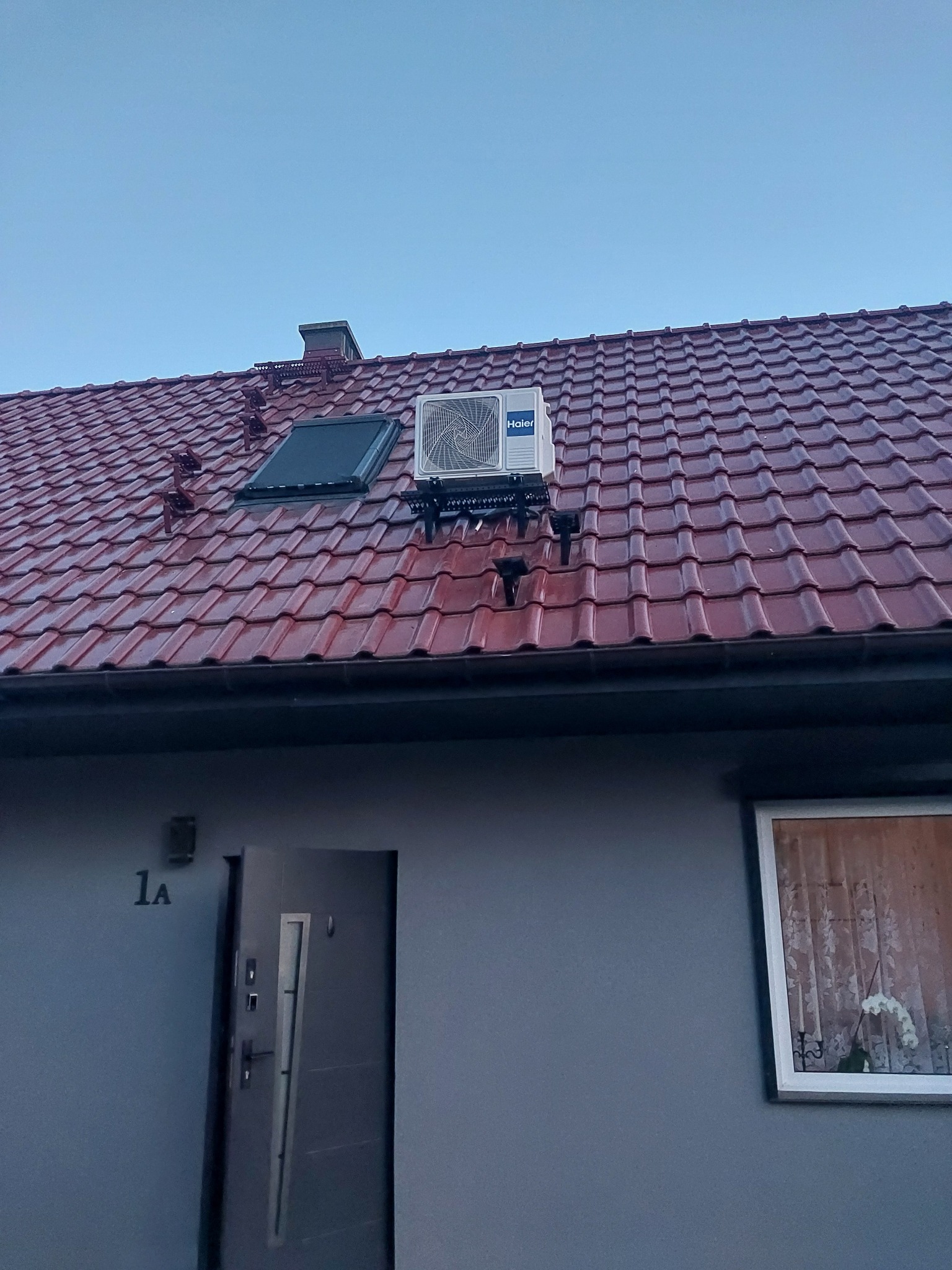 Biała jednostka zewnętrzna klimatyzatora Haier zamontowana na metalowym stelażu na czerwonym, błyszczącym dachu skośnym domu jednorodzinnego z oknem dachowym.
