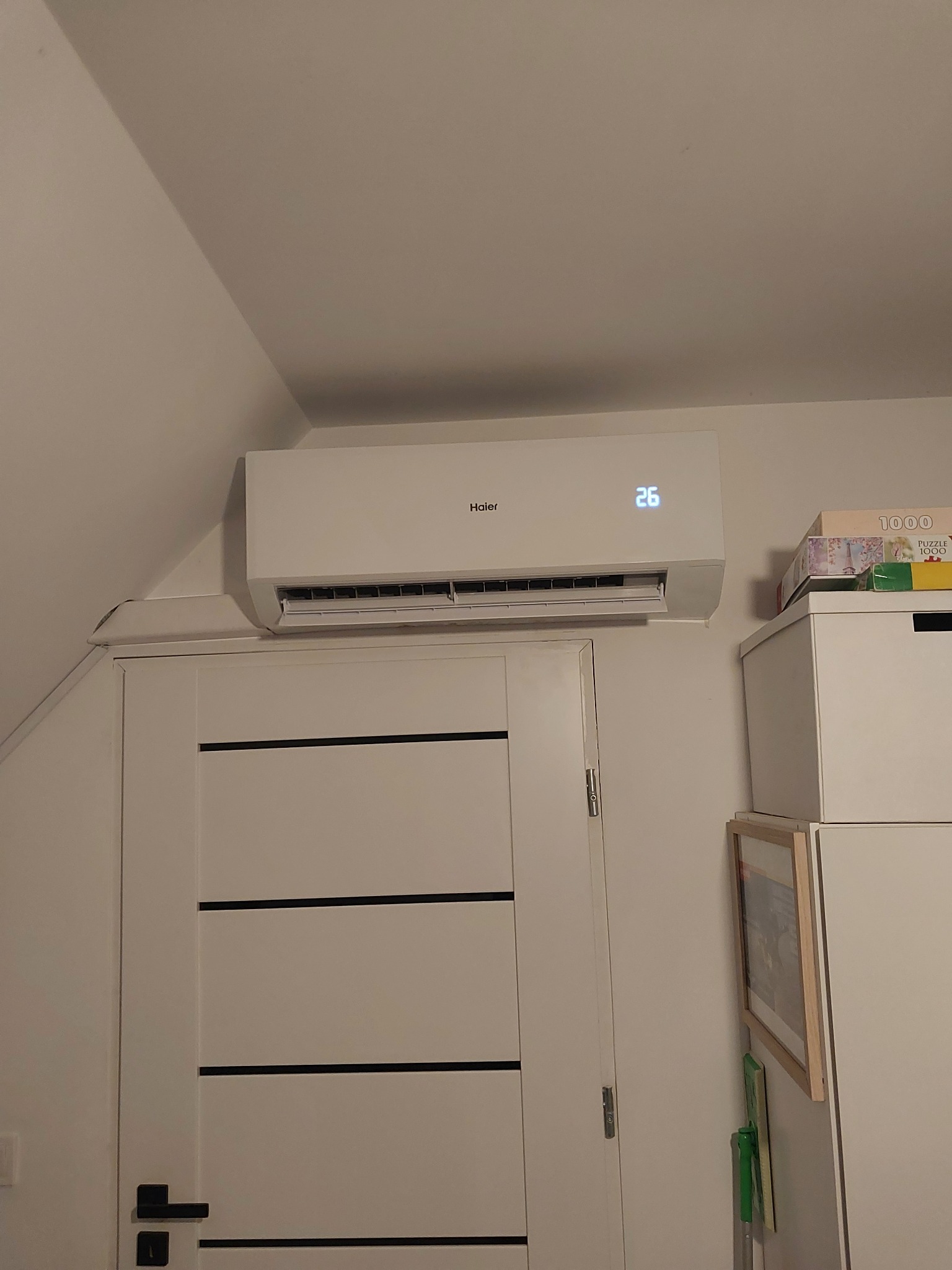 Biały klimatyzator Haier z wyświetlaczem temperatury 26 stopni Celsjusza zamontowany nad białą, nowoczesną drzwiami w pokoju na poddaszu.